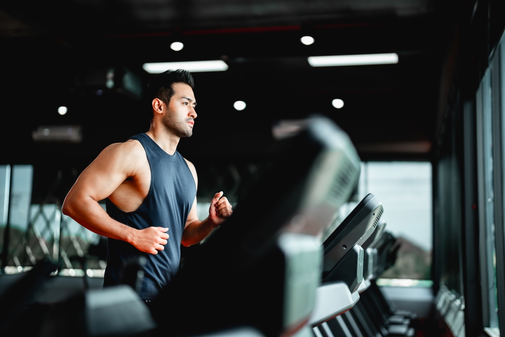 : "A-man-running-on-a-treadmill-in-a-gym-wearing-a-sleeveless-shirt-and-focusing-on-his-workout-with-views-of-the-gym's-windows-in-the-background.