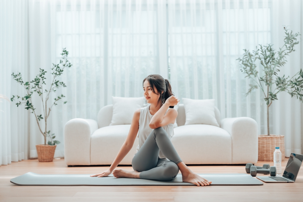 : "A-woman-doing-a-stretching-exercise-on-a-yoga-mat-at-home-she-is-wearing-gray-leggings-and-a-tank-top-and-sitting-cross-legged-with-her-hand-on-her-neck.