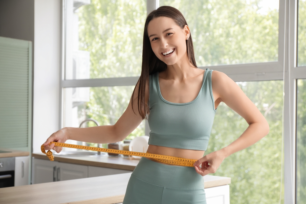 Woman-smiling-while-holding-measuring-tape-around-her-waist-in-a-bright-indoor-setting.