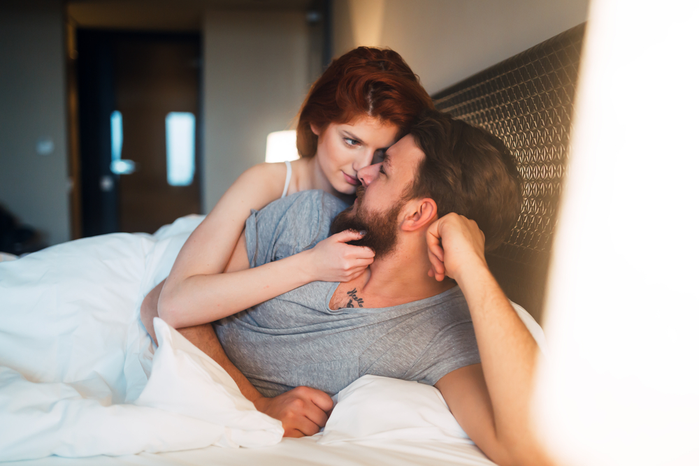 A-couple-in-bed-sharing-a-tender-moment-the-woman-is-holding-the-man's-neck-while-gazing-at-him-the-man-smiles-as-the-woman-leans-in-close.