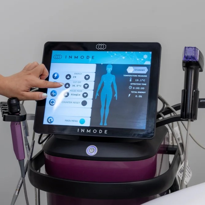 A-hand-touching-the-touchscreen-of-an-INMODE-device-with-settings-for-energy-level-pulse-mode-temperature-measurement-and-total-energy-the-device-is-purple-and-connected-to-cables.