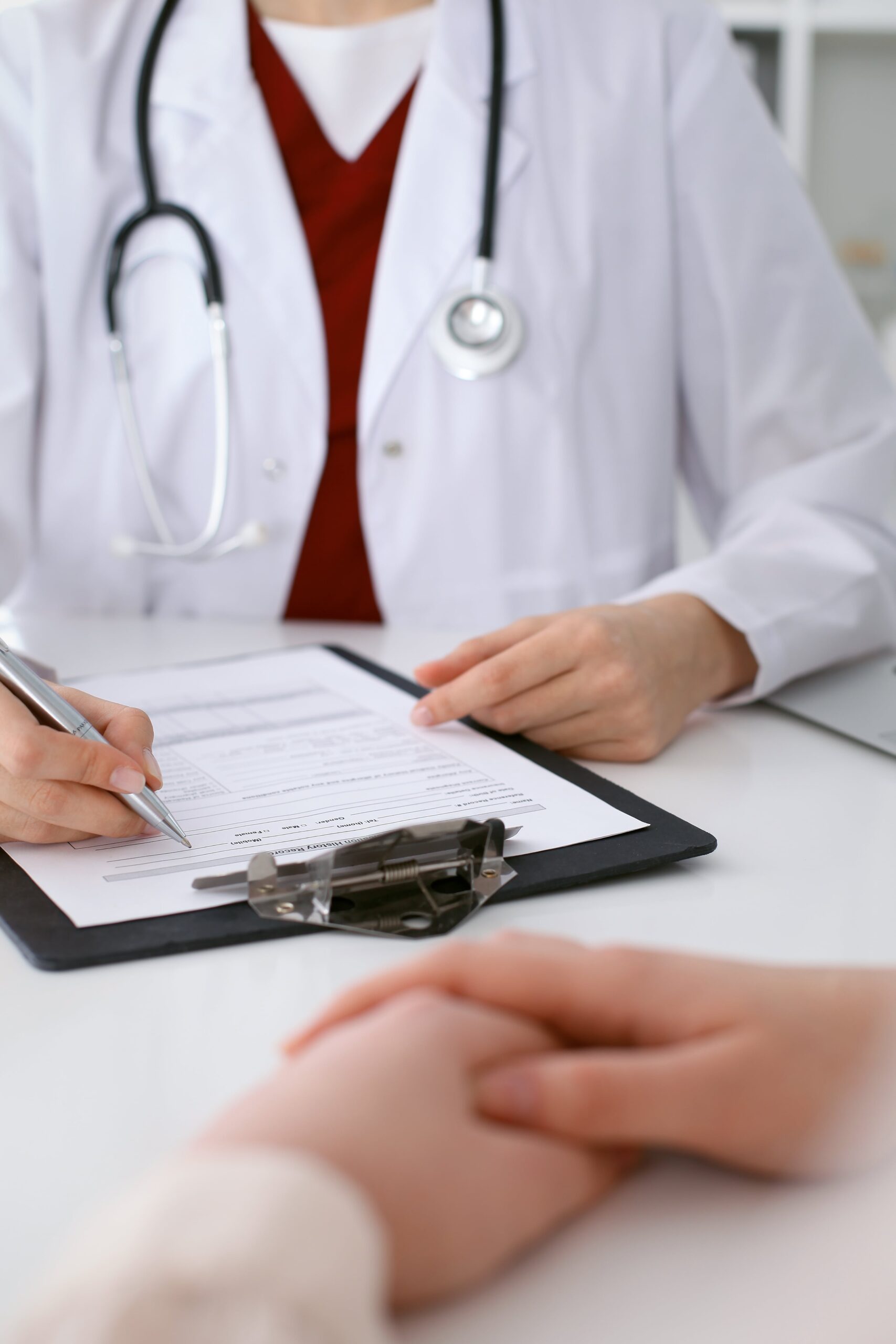 : "A-doctor-in-a-white-coat-with-a-stethoscope-is-writing-on-a-medical-form-with-a-pen-A-person's-hand-is-resting-on-the-table-in-front-of-them-the-form-is-secured-with-a-clipboard.