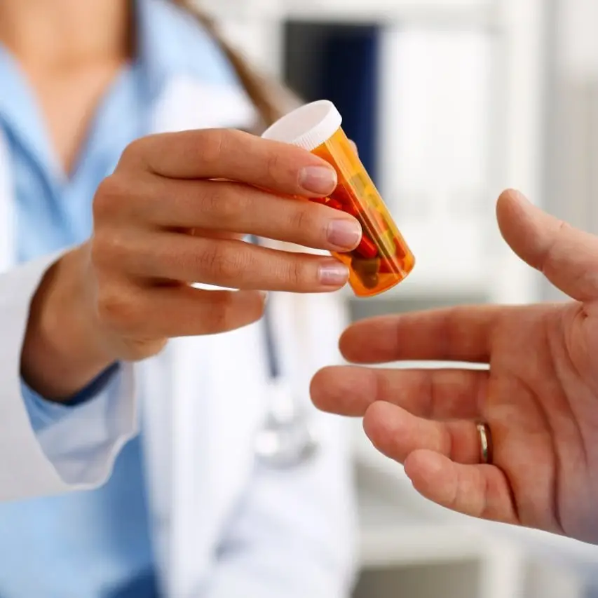 A healthcare professional in a white coat handing an orange prescription bottle with pills to a patient.